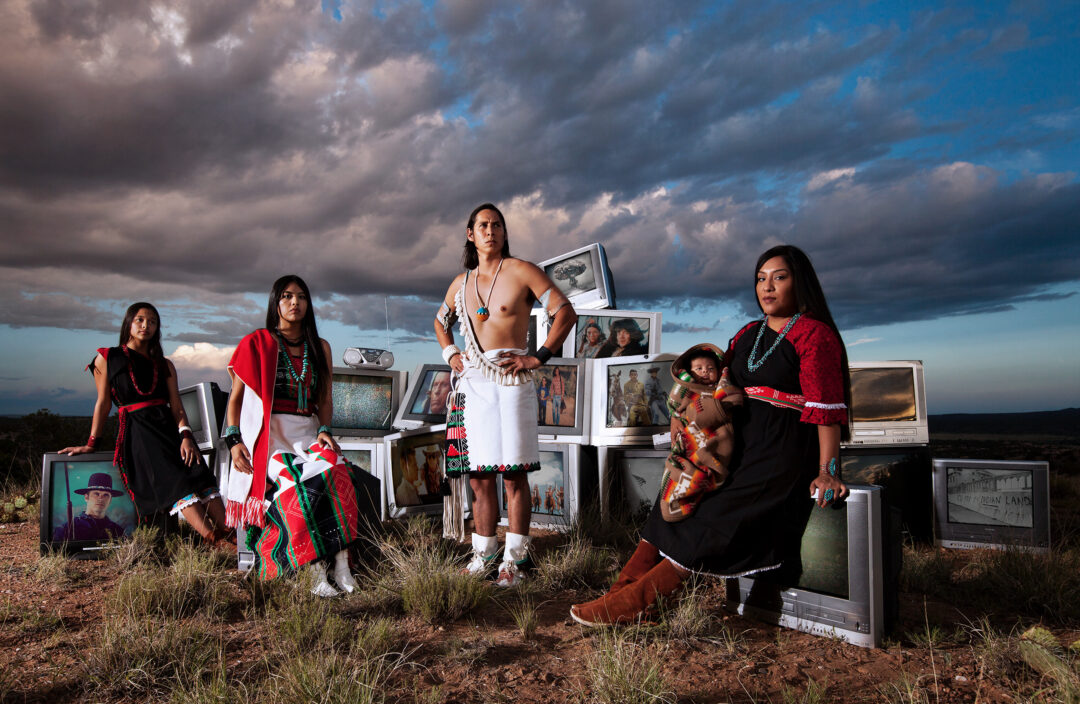 A group of Native Americans outdoors standing and sitting in front of old tvs